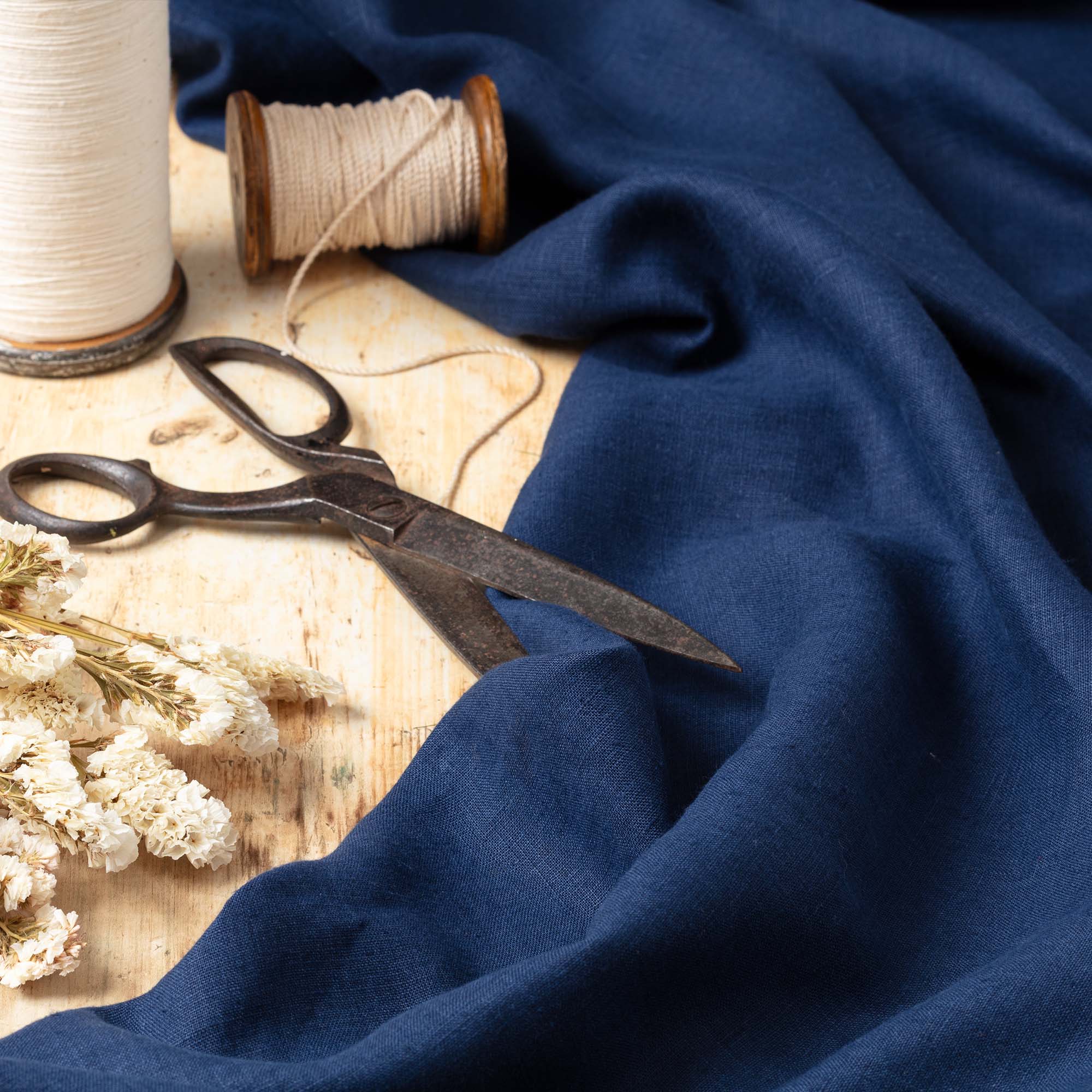 Dark Blue linen fabric on wooden surface, ready to cut, with scissors, thread bobbins and flowers