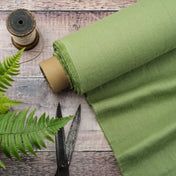 Fern Green linen fabric on wooden table, ready to cut, with scissors, thread bobbins and fern plant