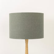 A dusty sage lampshade on a wooden lamp base, with a textured weave design, photographed against a white background.