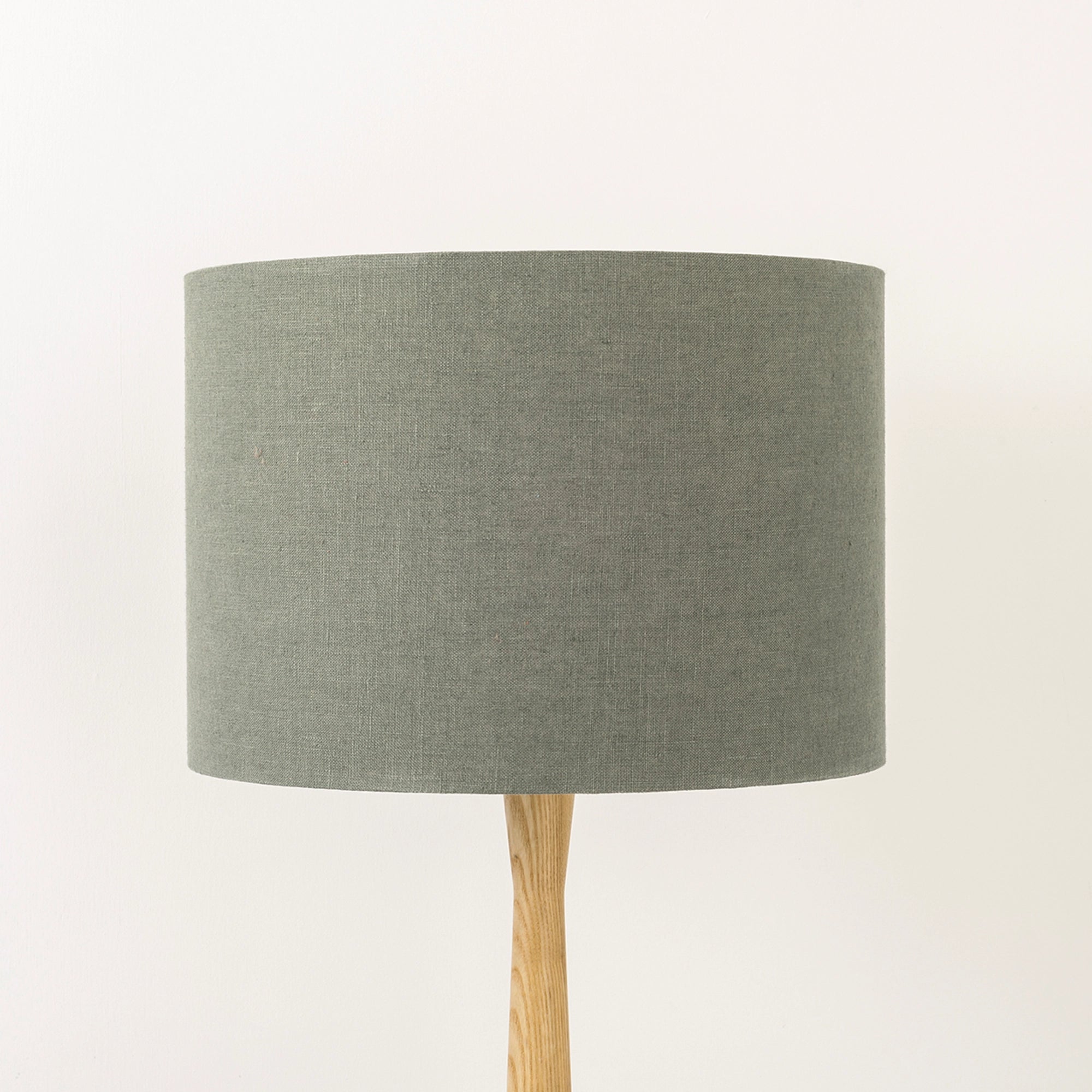 A dusty sage lampshade on a wooden lamp base, with a textured weave design, photographed against a white background.