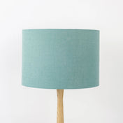 A dusty turquoise linen lampshade on a wooden lamp base, with a textured weave design, photographed against a white background.
