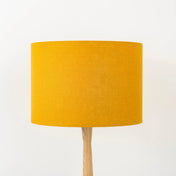 A mustard yellow lampshade on a wooden lamp base, with a textured weave design, photographed against a white background.
