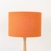 A pumpkin orange linen lampshade on a wooden lamp base, with a textured weave design, photographed against a white background.