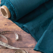 Deep teal linen fabric on the roll, unrolled beside a tailor's measuring tape on a wooden table